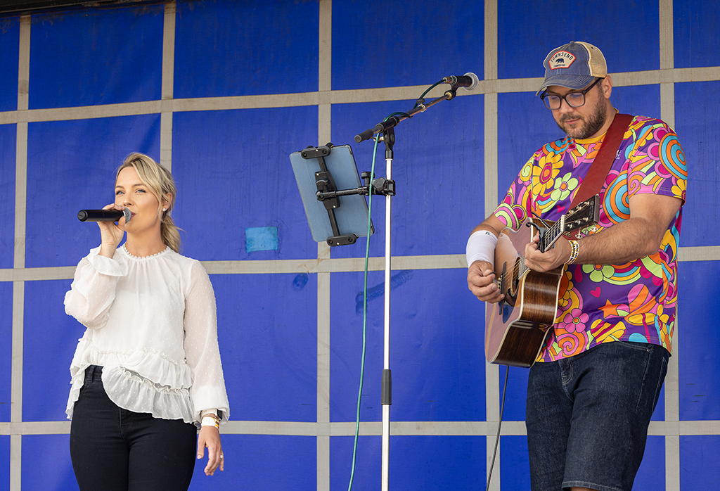 A live musical duo performing on a stage with a gridded blue background, featuring a female vocalist singing into a microphone on the left and a male guitarist wearing a bright psychedelic shirt on the right.